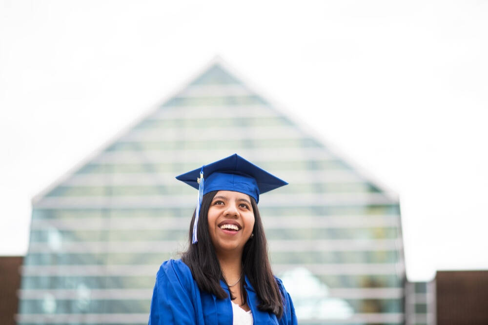 A young woman wearing a blue graduation cap and gown smiles as she stands in front of a modern glass‑and‑steel building that appears campus‑like.
